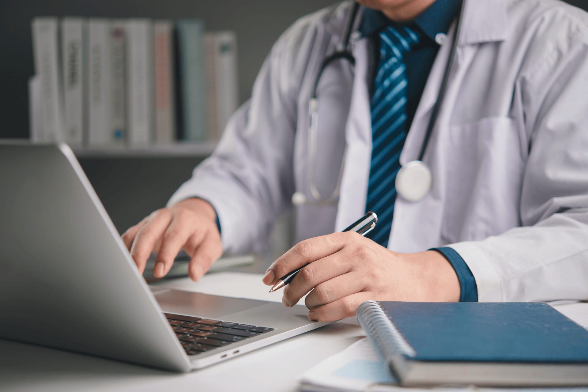 A doctor working on a laptop with a notebook on the desk.