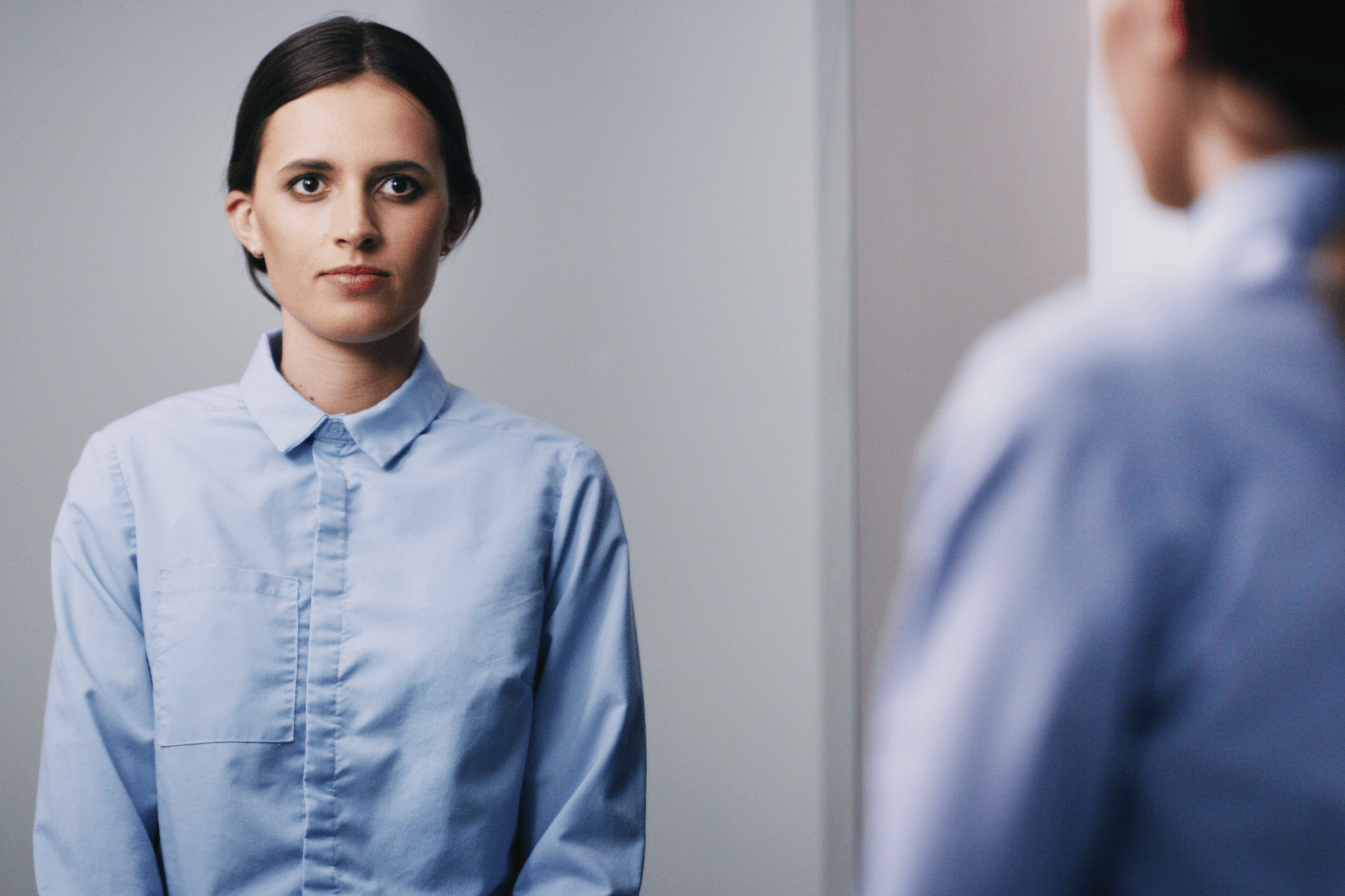 Woman in a blue shirt looking at herself in a mirror with a serious expression.