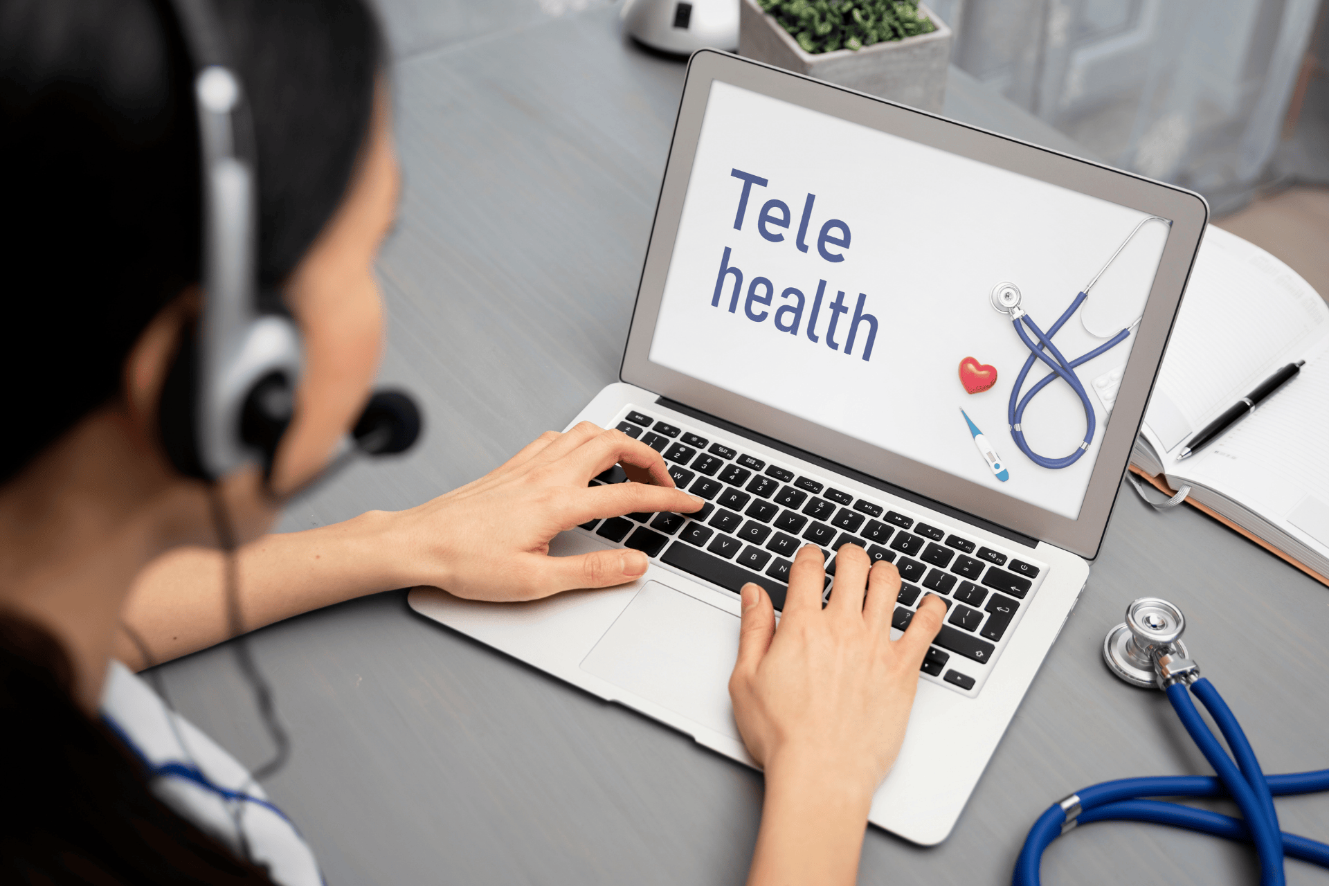 Doctor wearing a headset typing on a laptop with a telehealth screen.