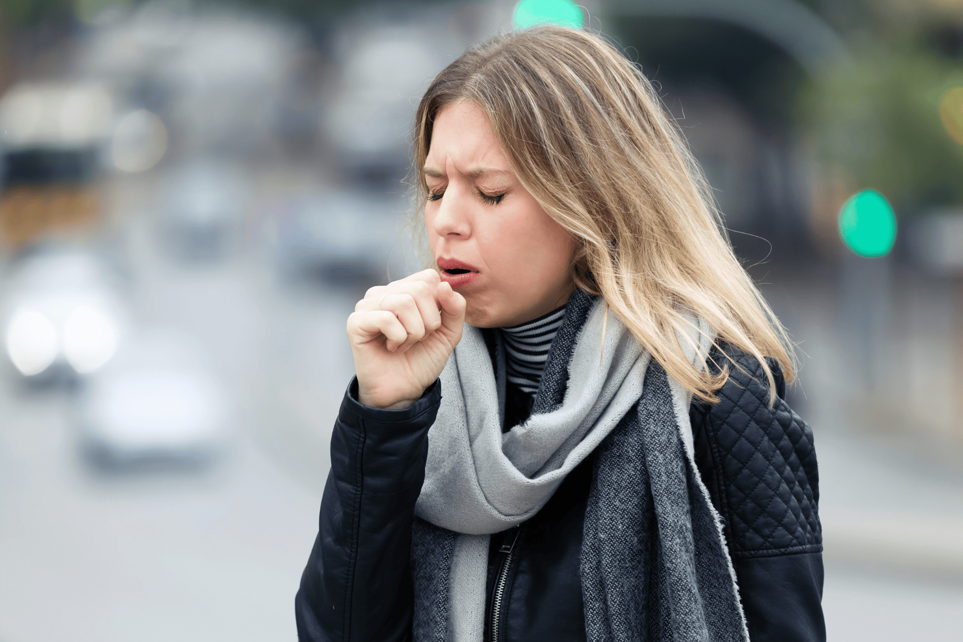 Woman coughing outdoors while wearing a scarf and jacket.