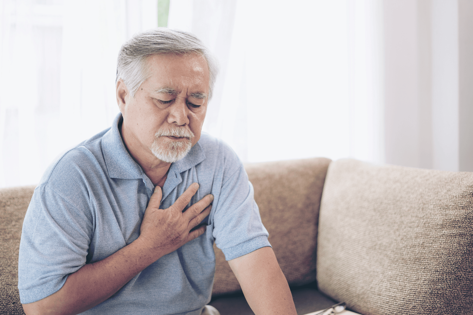 Elderly man sitting on a couch holding his chest in discomfort.