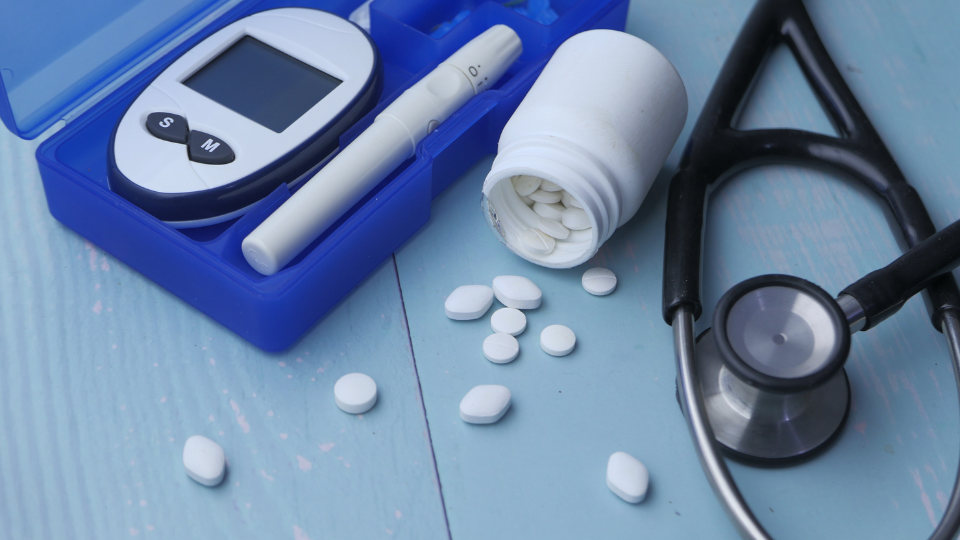 Glucose checker, stethoscope, and bottle of pills spilling out on a blue table