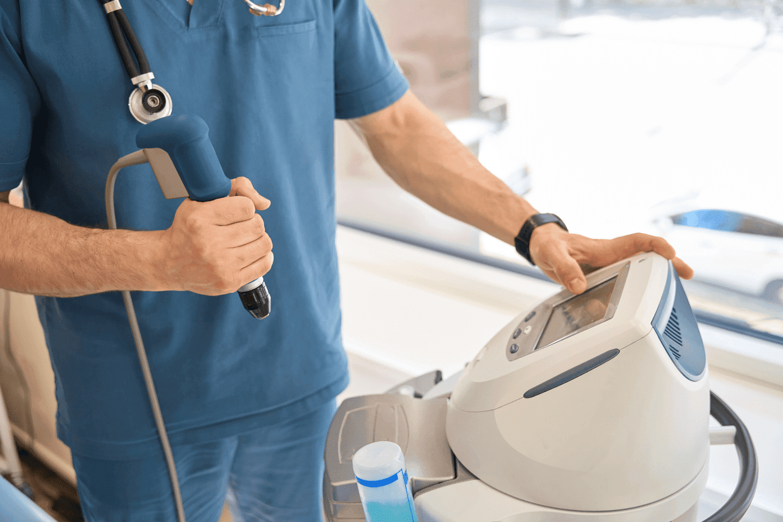 Healthcare worker in blue scrubs holding a medical device beside a clinic machine.