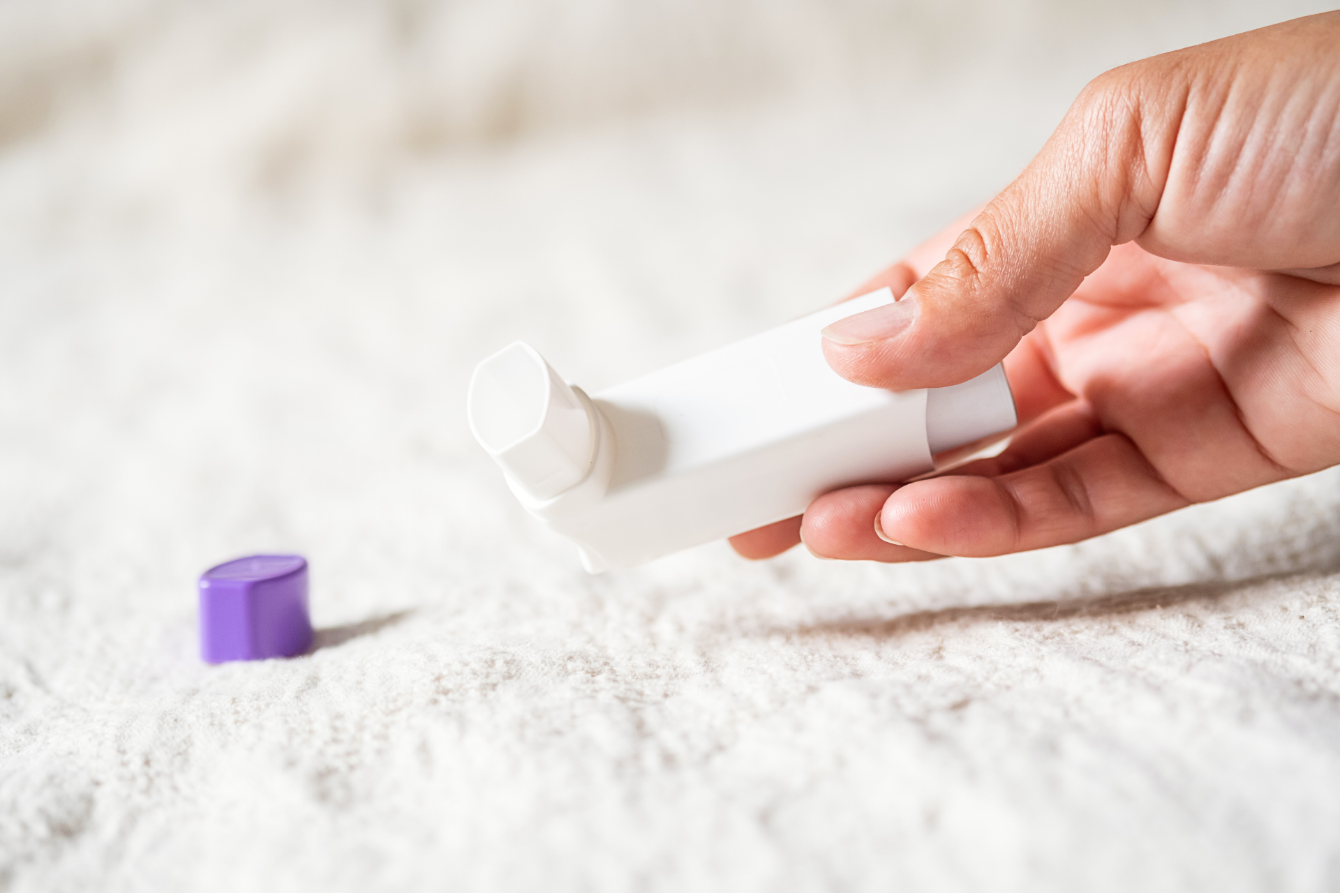 Hand holding a white asthma inhaler with its purple cap lying nearby
