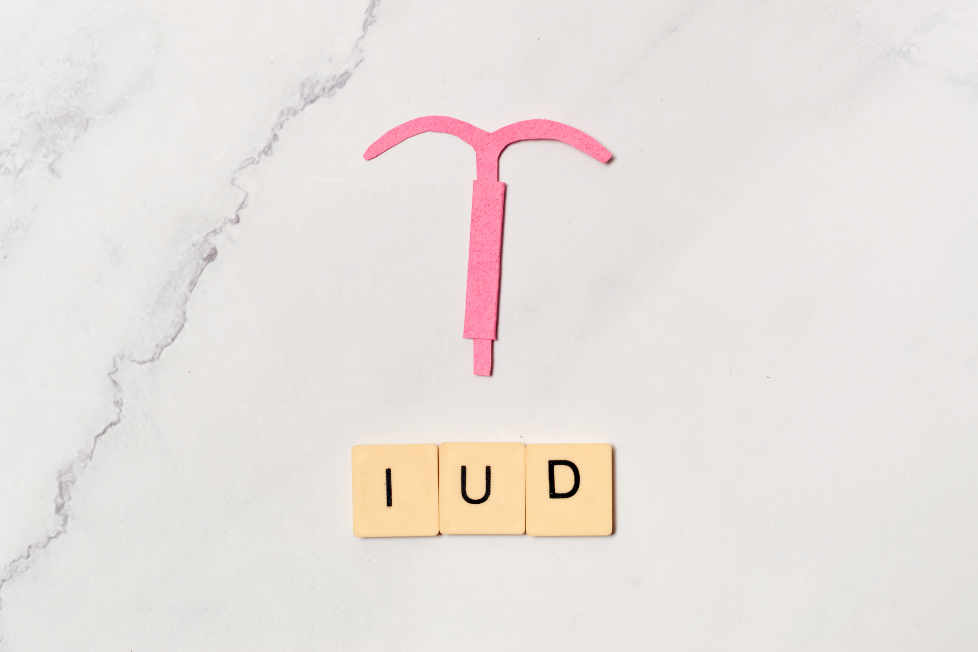 Pink cutout of an IUD above letter tiles spelling ‘IUD’ on a marble surface