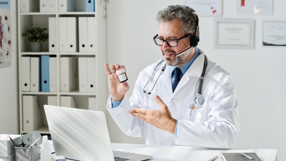 Male doctor on laptop for telehealth video call holding up a pill bottle.