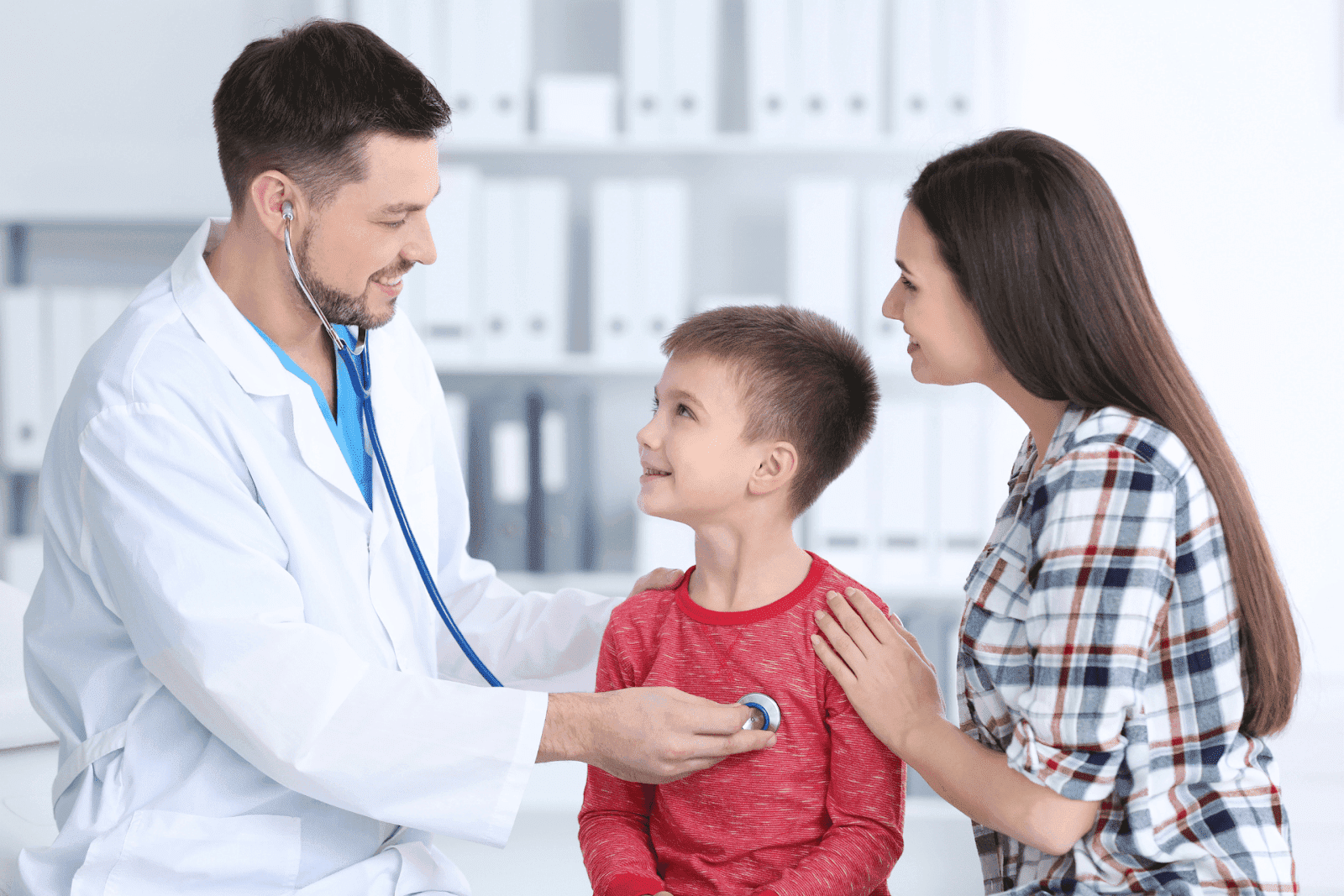 Male doctor using a stethoscope to examine a smiling young boy, with the boy’s mother beside him