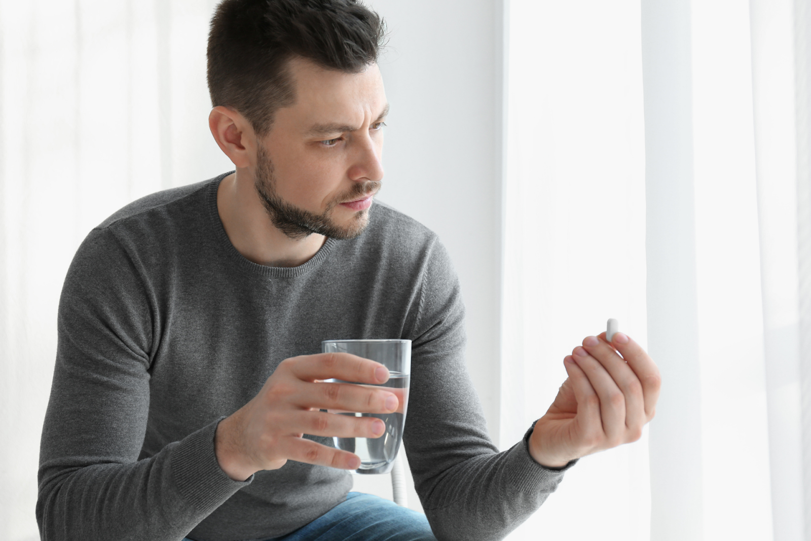 Man holding a glass of water and looking at a pill in his other hand
