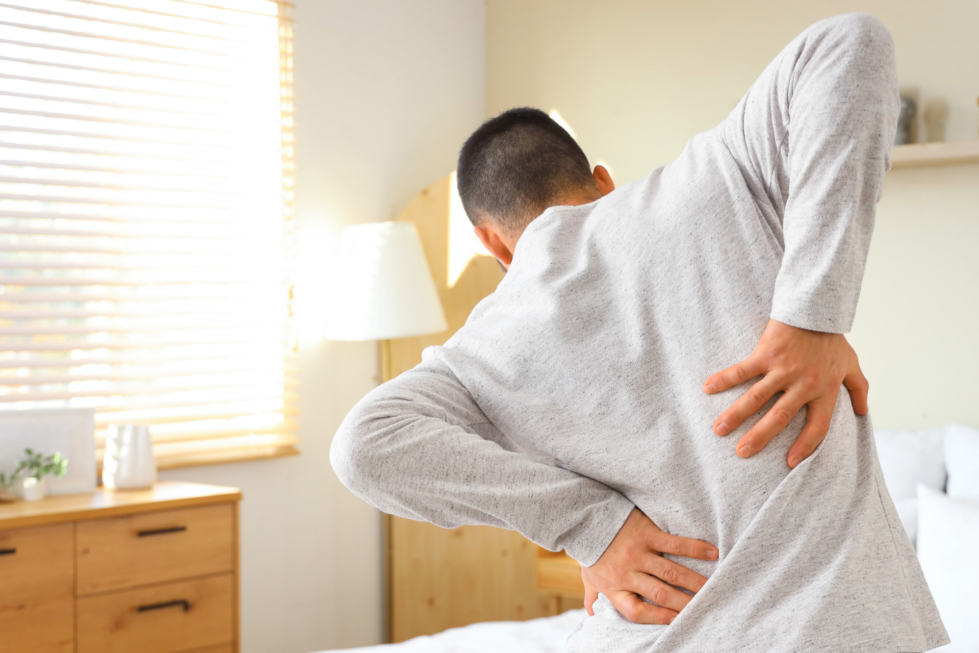 Man holding his lower back in pain while standing in a bedroom