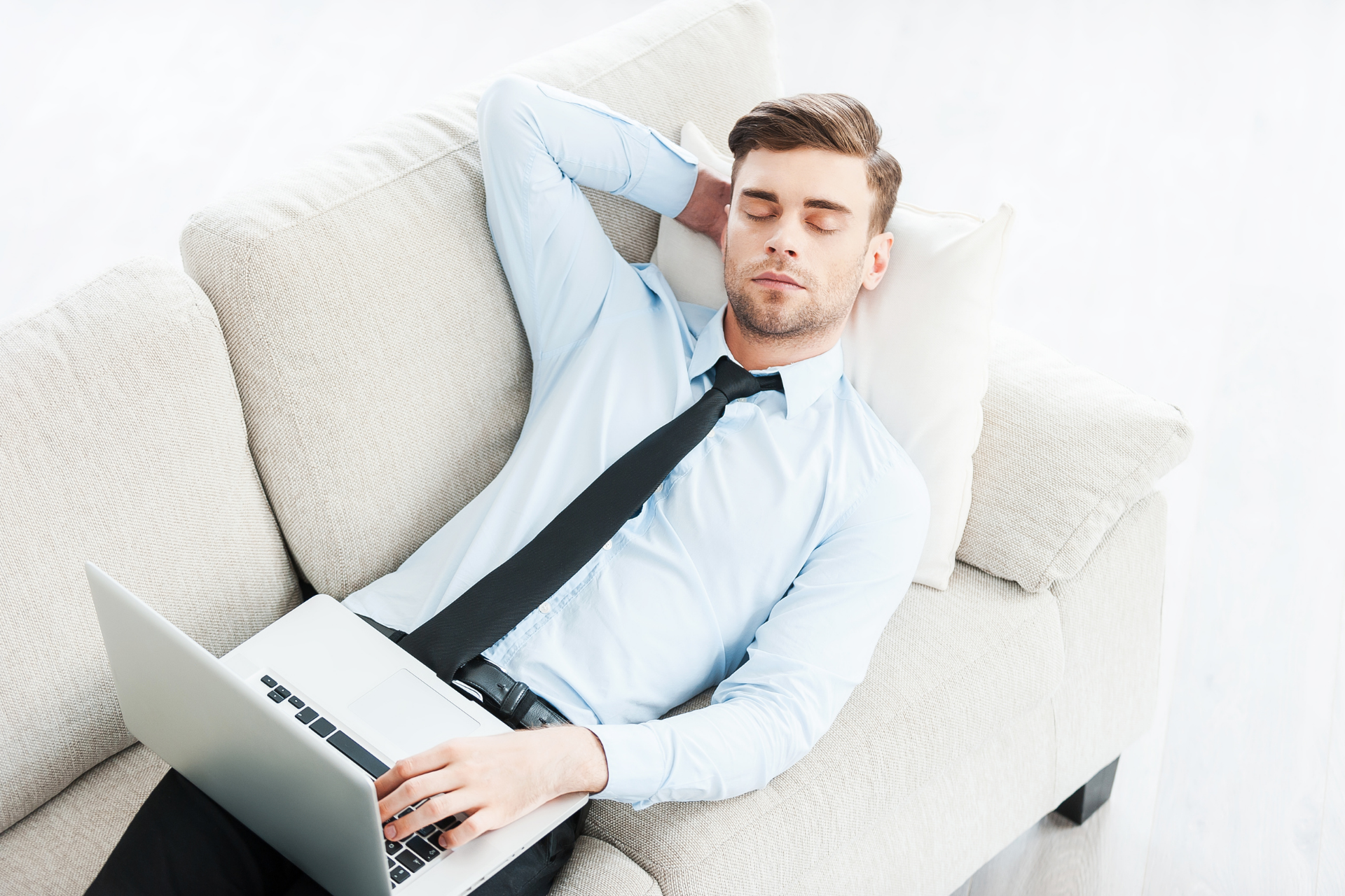 Man in business attire resting on a couch with eyes closed, laptop on his lap, appearing tired or unwell at home