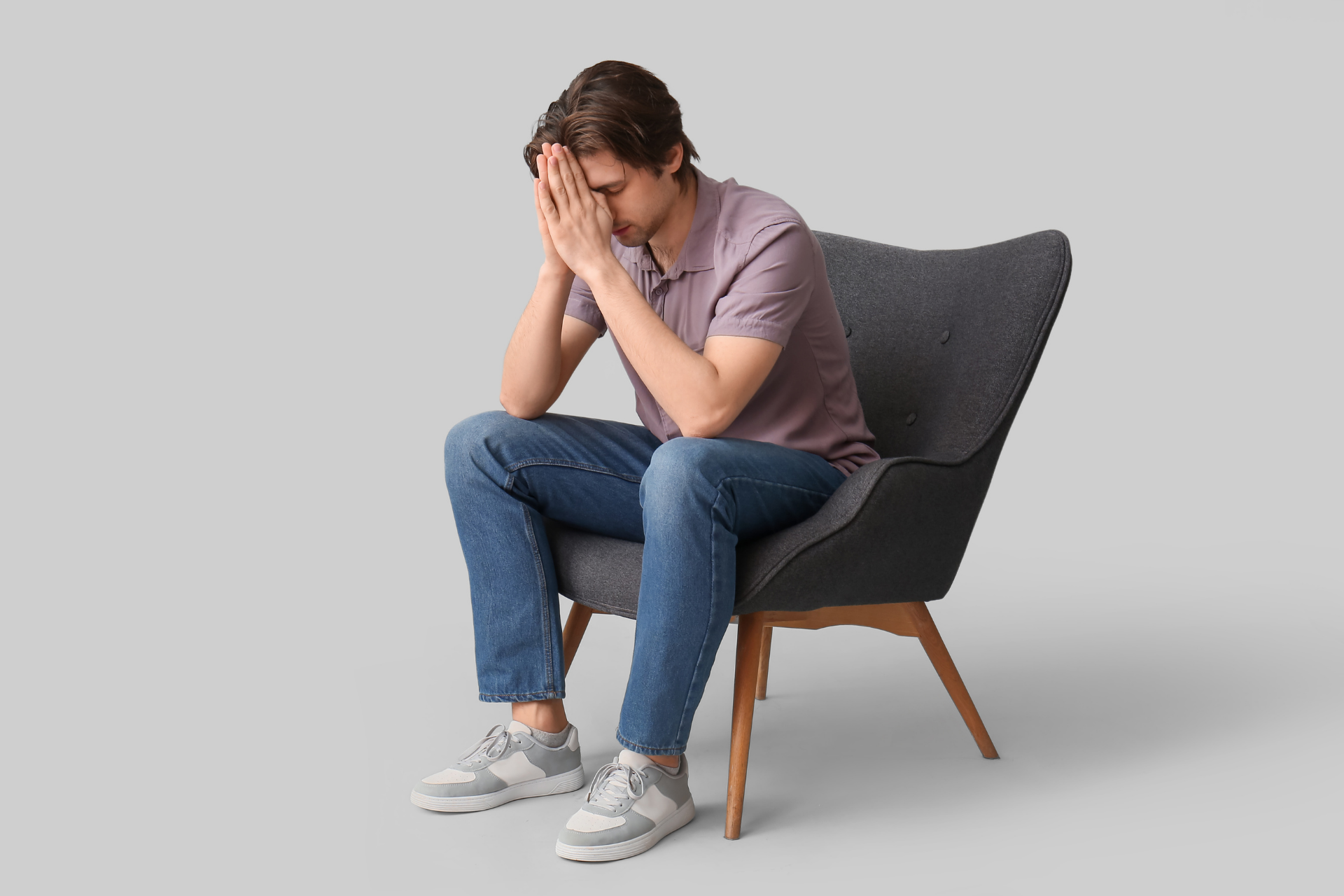 Man sitting on a chair leaning forward with hands pressed to his face, appearing stressed or unwell against a light background
