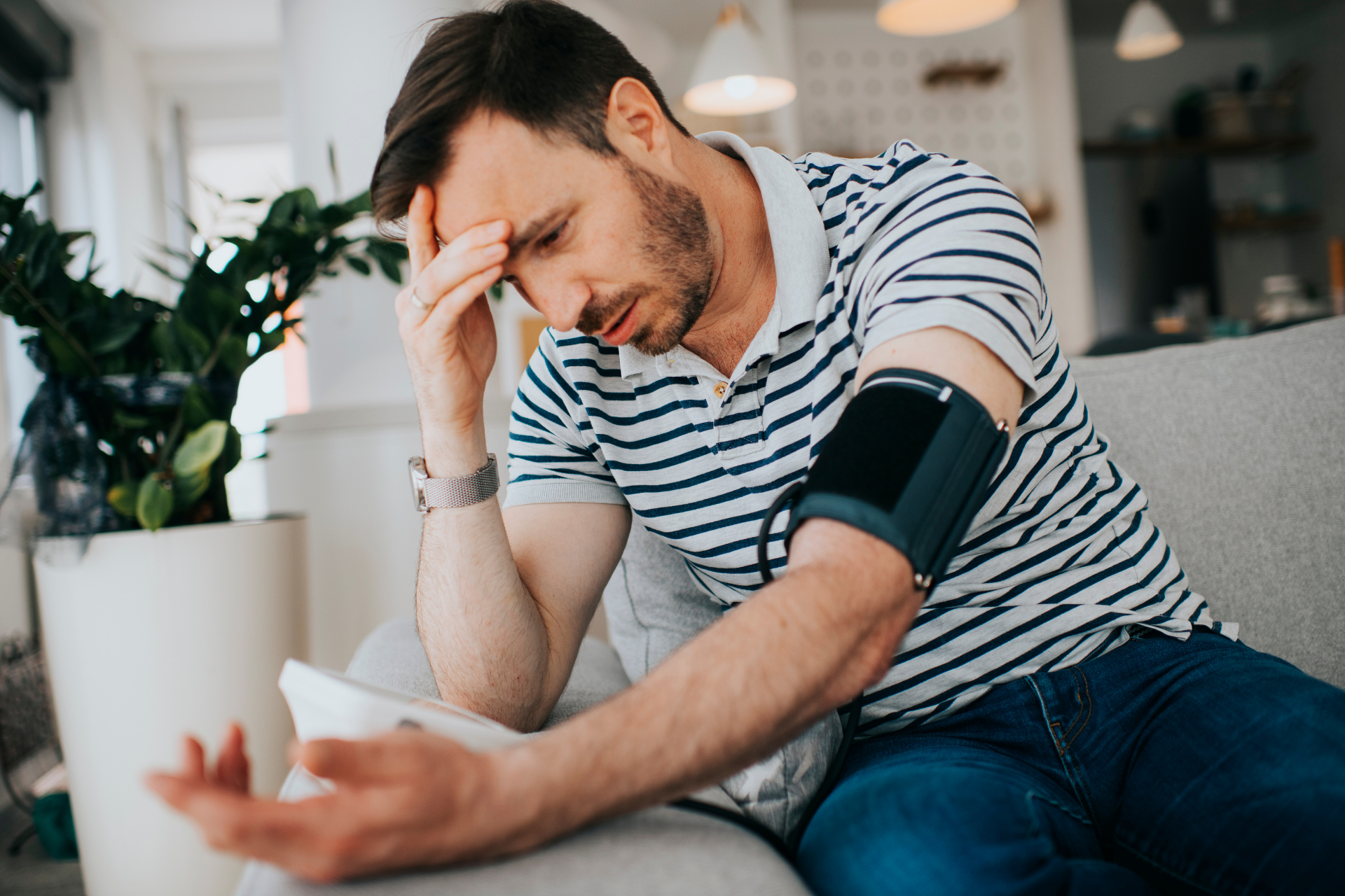 Man sitting on a couch checking his blood pressure at home, holding his head and looking concerned