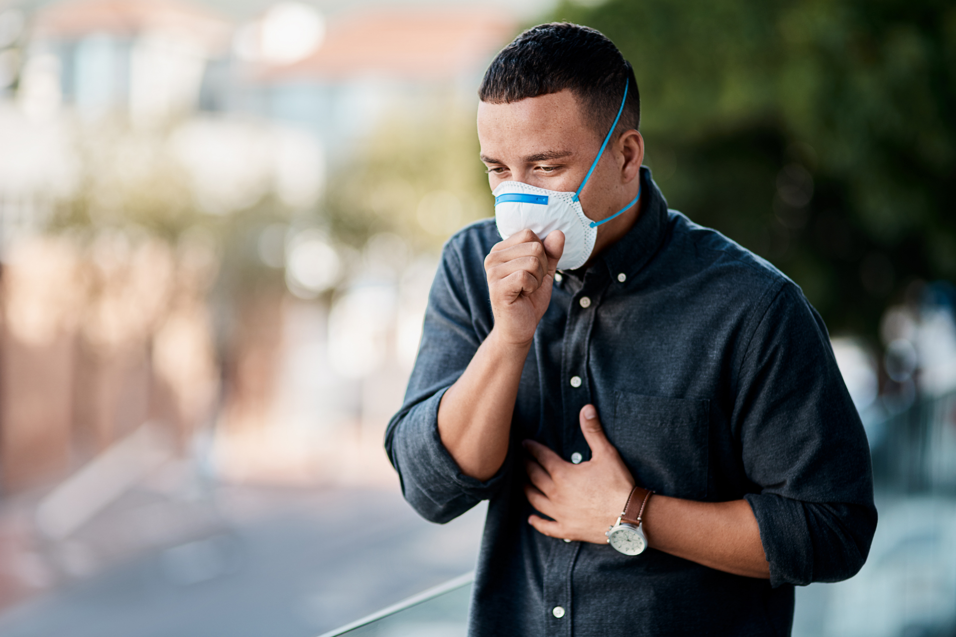 Man wearing a face mask and coughing while standing outdoors, holding his chest