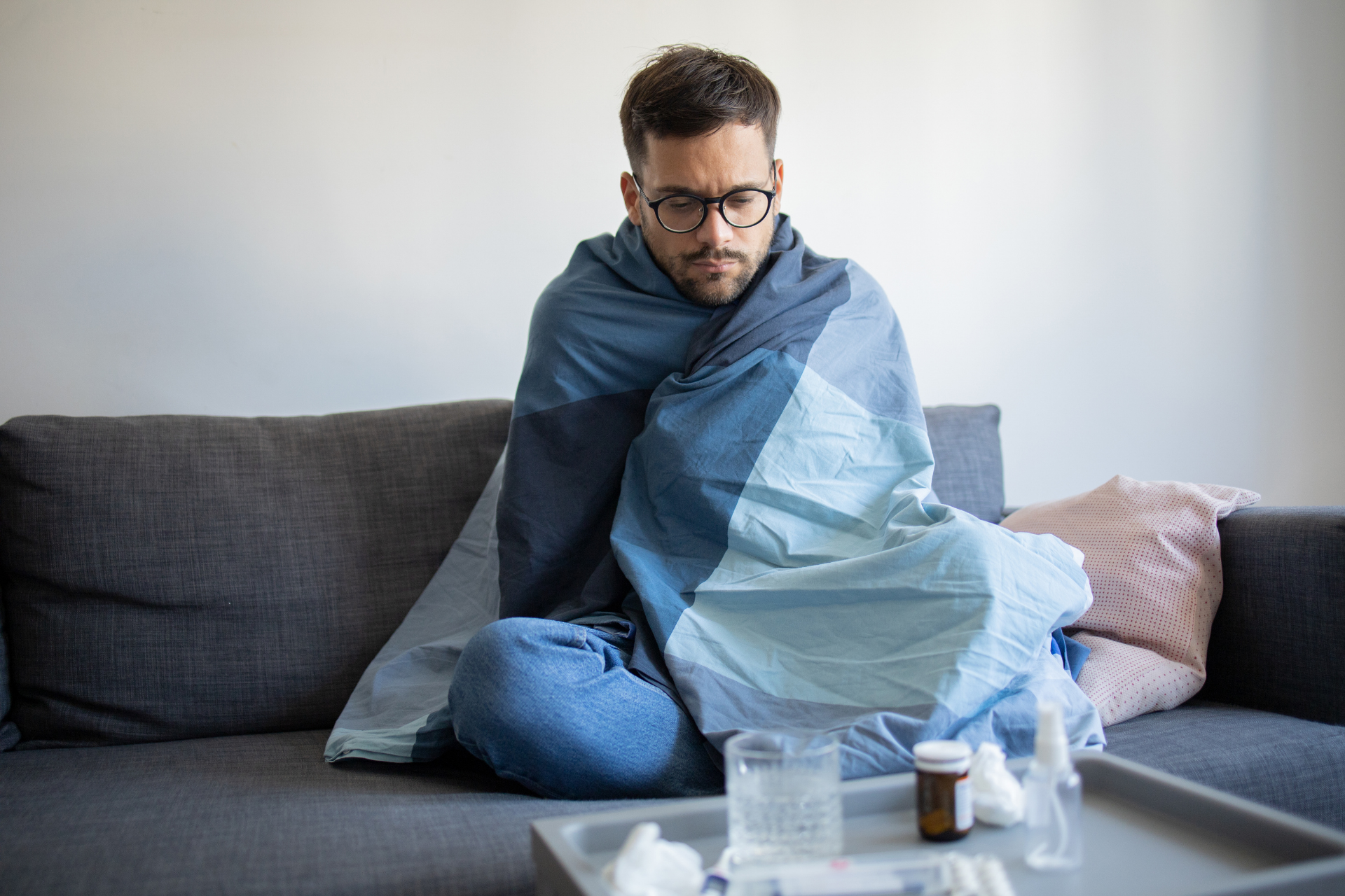 Man wrapped in a blanket sitting on a couch with medicine and tissues, appearing unwell
