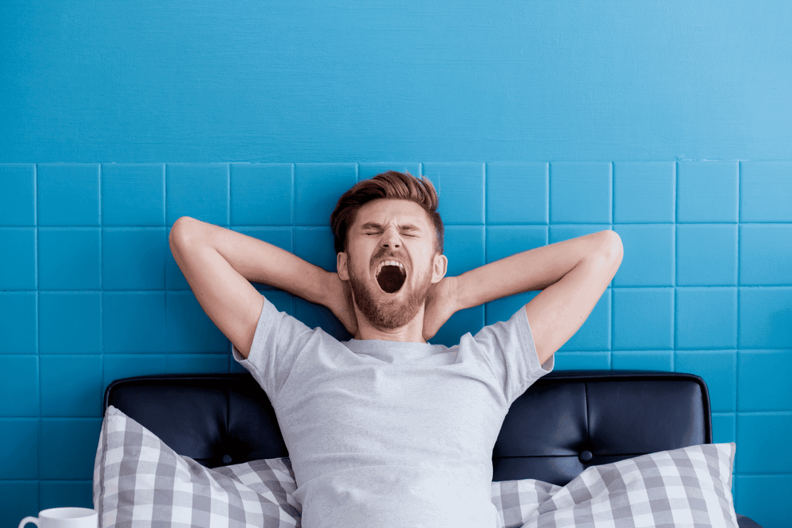 Man yawning while stretching in bed against a blue wall.