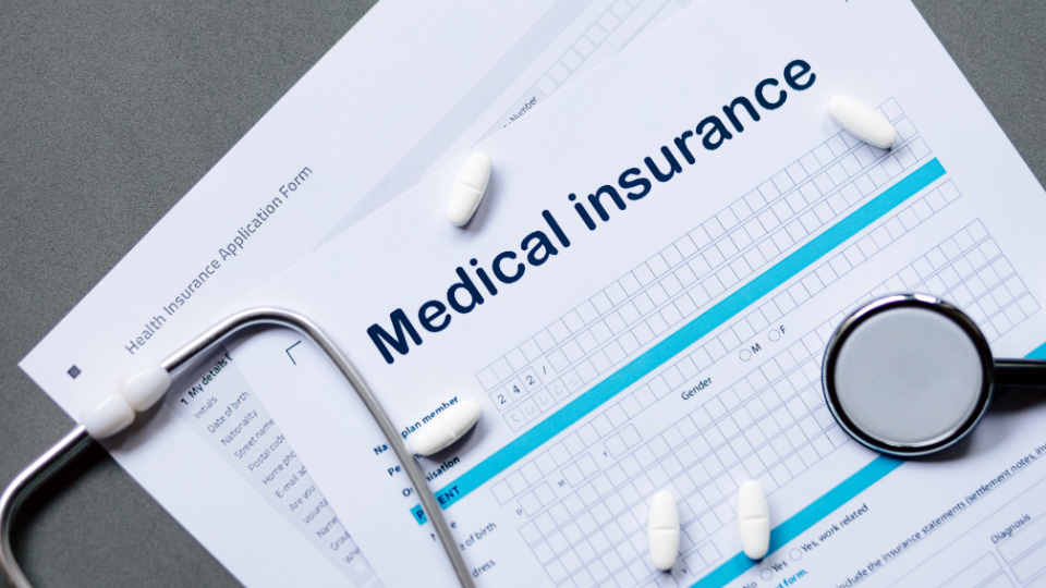 Medical insurance application forms with some pills and a stethscope on top of them.