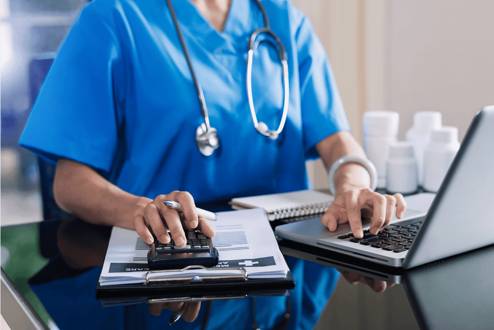 Medical professional in blue scrubs using a calculator and laptop at a desk.