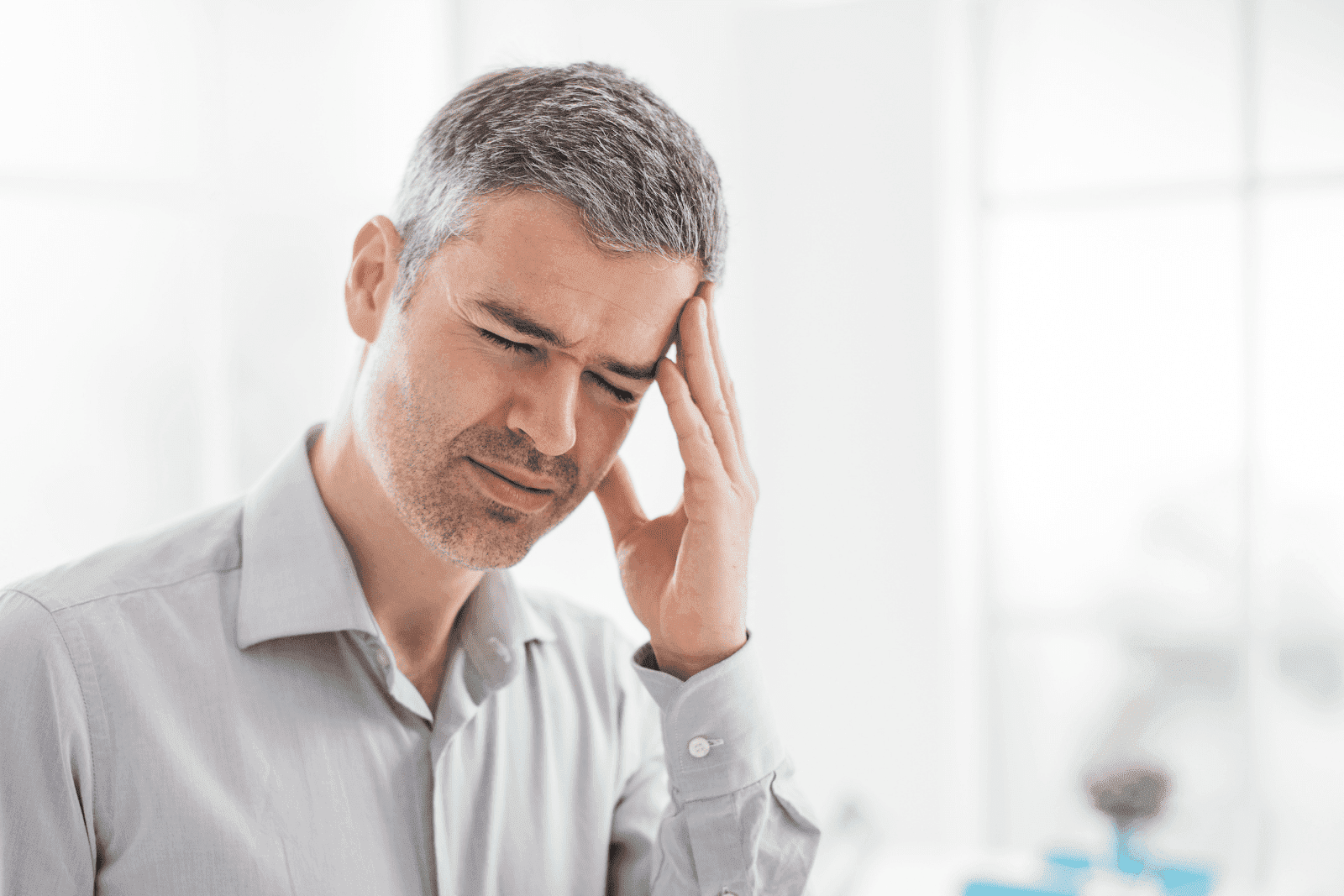Middle-aged man in a light shirt indoors, closing his eyes and holding his head with a pained expression