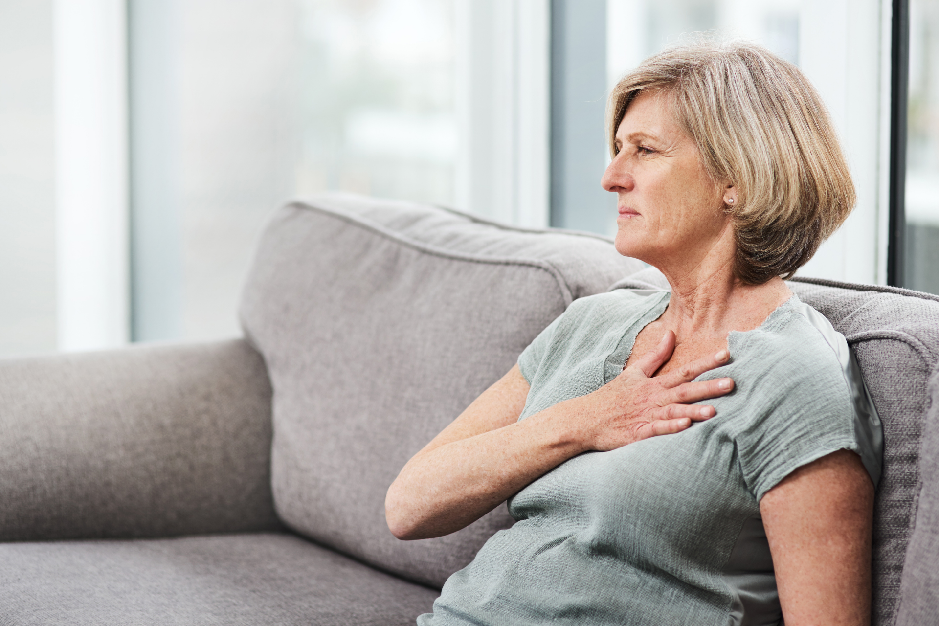 Middle-aged woman sitting on a couch holding her chest, appearing uncomfortable or concerned