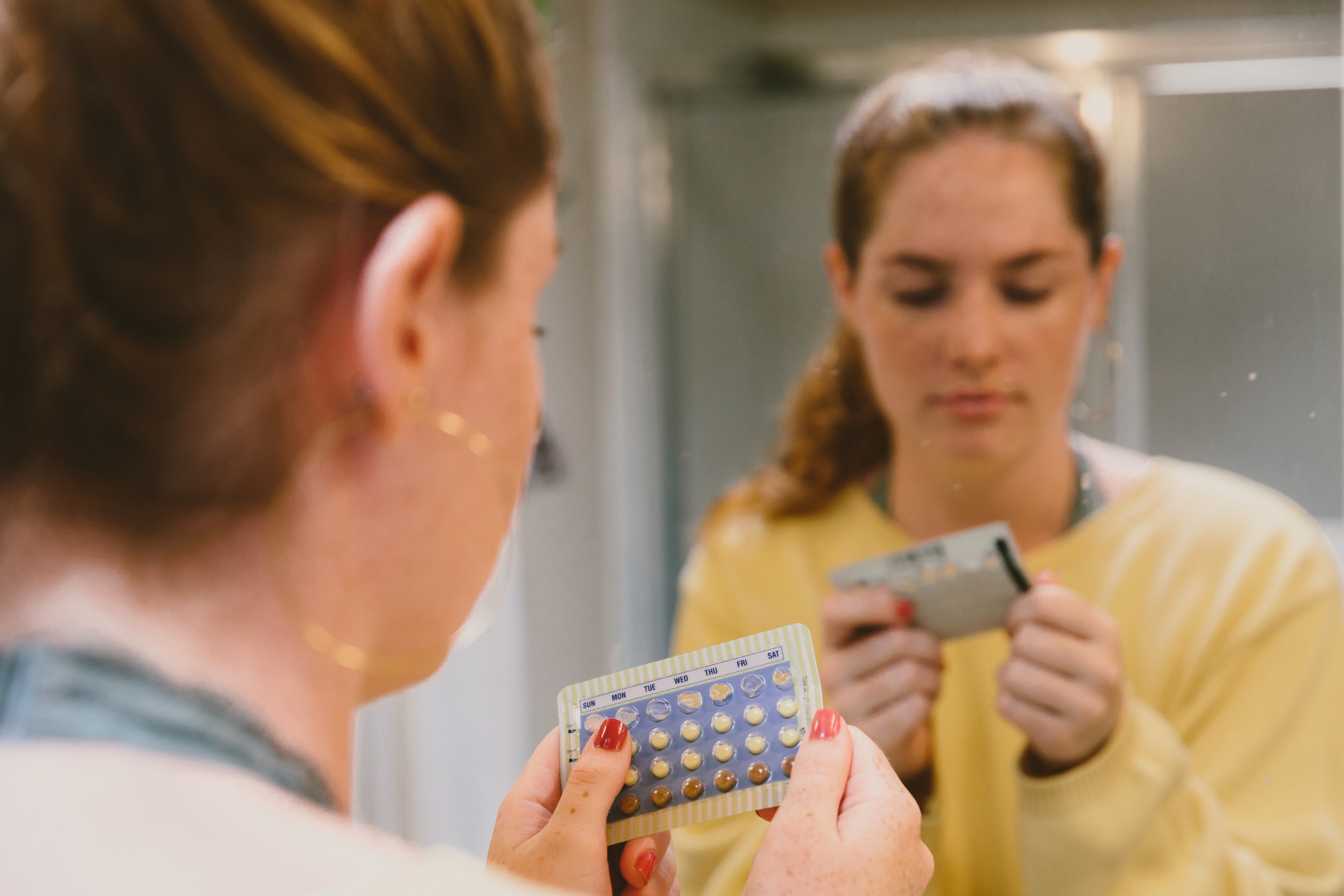 Woman looking at a pack of birth control pills in front of a bathroom mirror