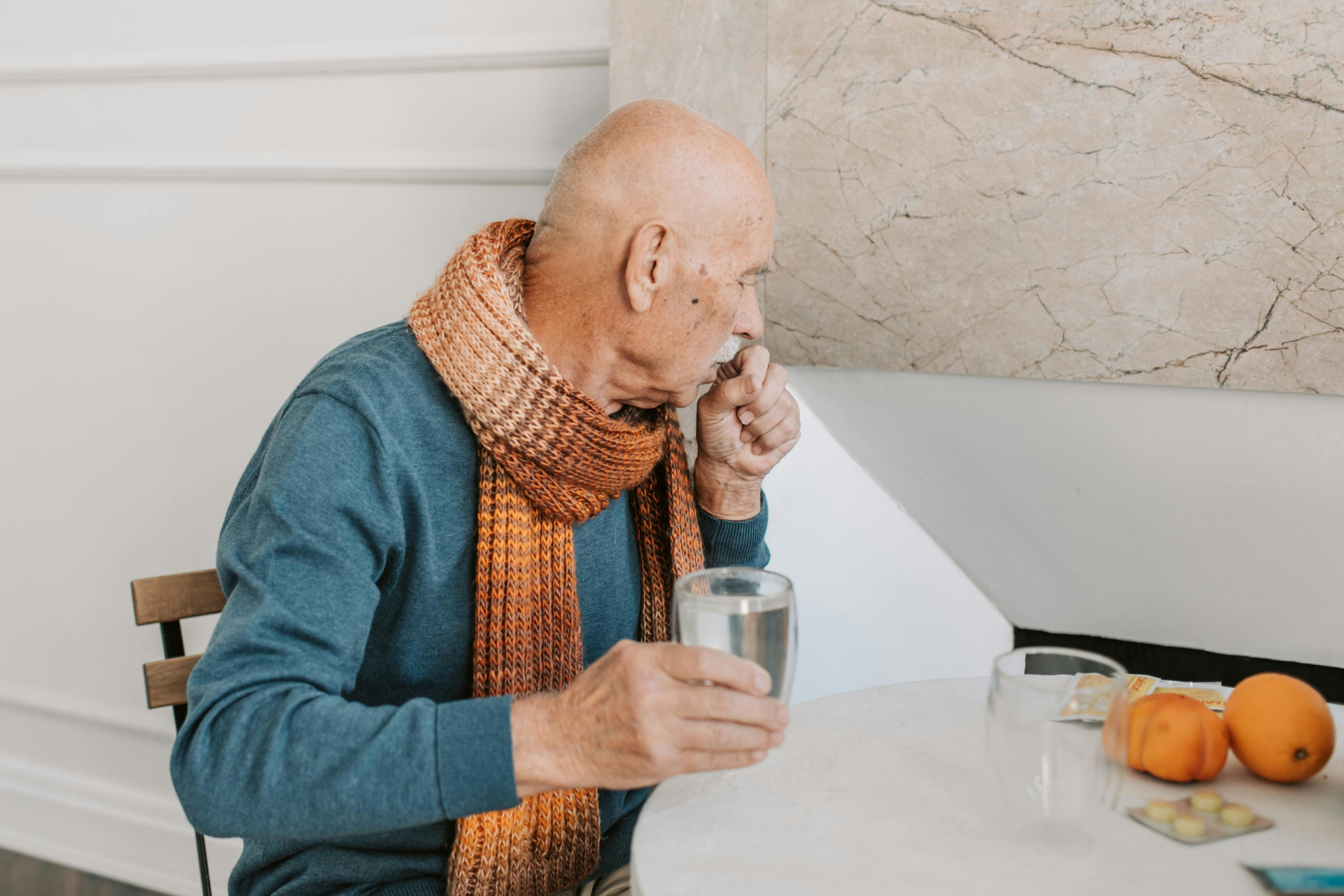 Older adult wearing a scarf, coughing while holding a glass of water at a table