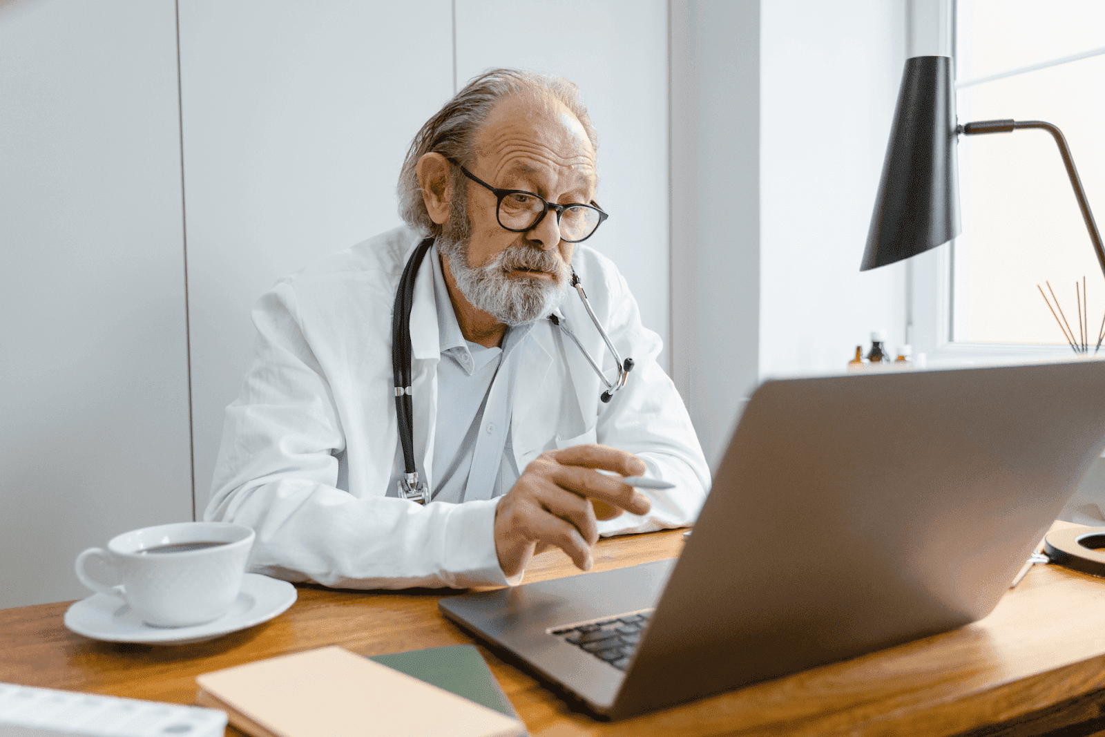 Older male doctor wearing glasses and a stethoscope working on a laptop at his desk