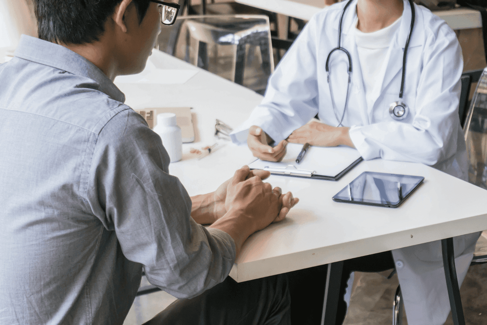 Patient consulting with a doctor in a white coat at a desk with a clipboard and tablet during a medical appointment.
