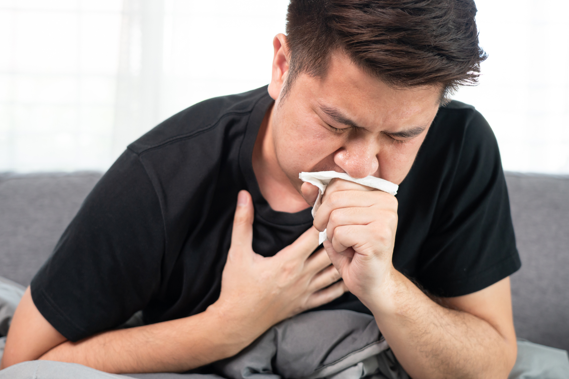 Person coughing into a tissue while holding their chest, sitting on a couch