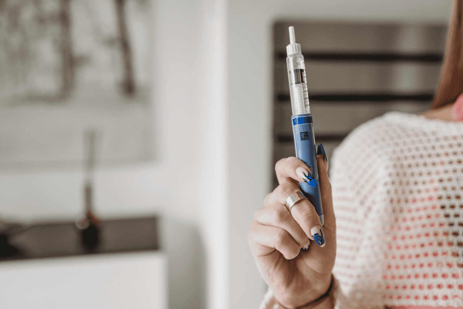 Person holding a blue insulin pen or injection pen with manicured nails and silver rings, in a bright indoor setting.