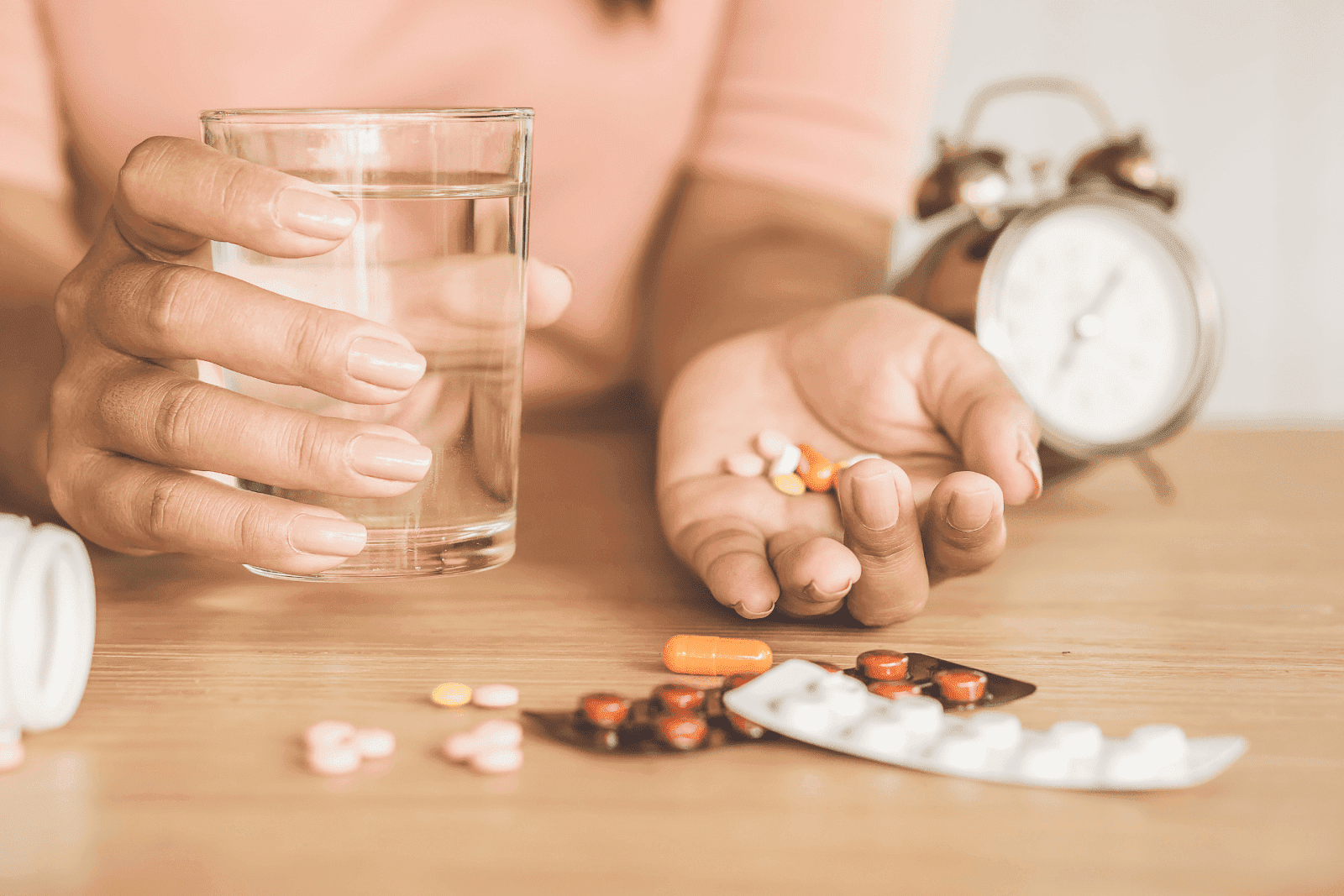 Person holding a glass of water in one hand and several pills in the other, preparing to take medication at a wooden table.