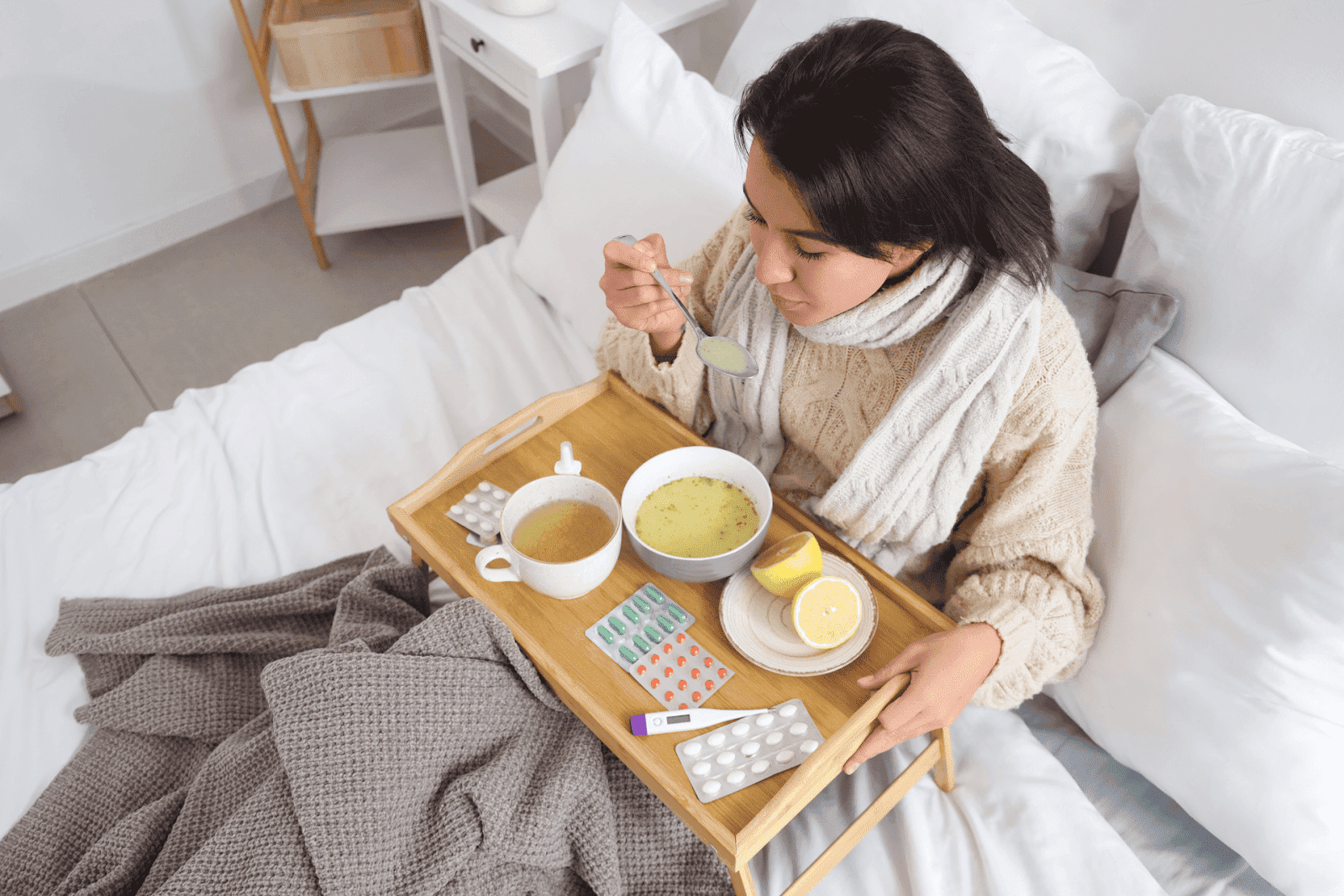 Person sitting in bed eating soup from a tray with medicine, tea, and lemon nearby