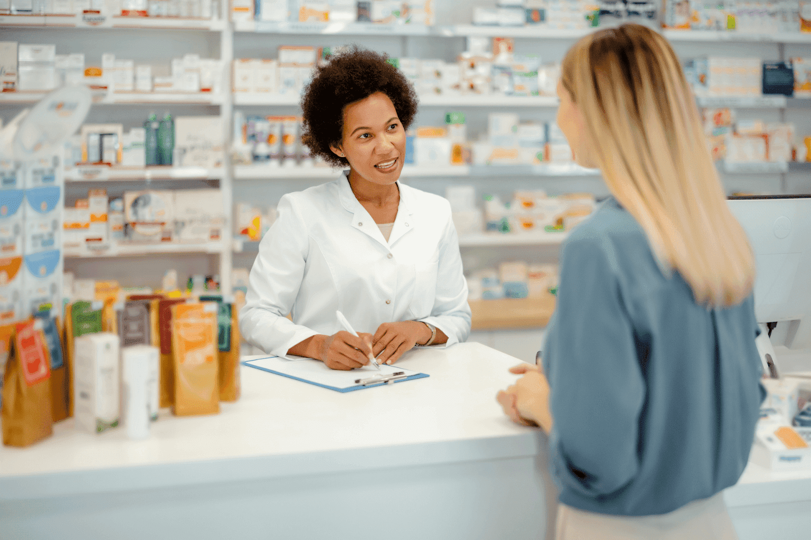 Pharmacist in a white coat talking with a customer at a pharmacy counter.