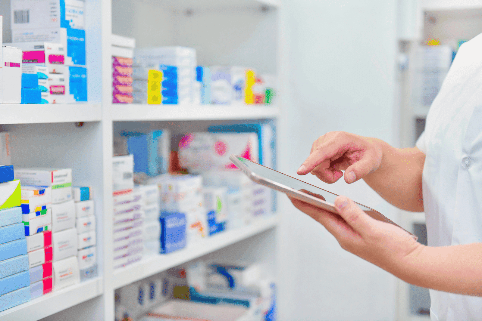 Pharmacist using a tablet in front of shelves stocked with medicine boxes.