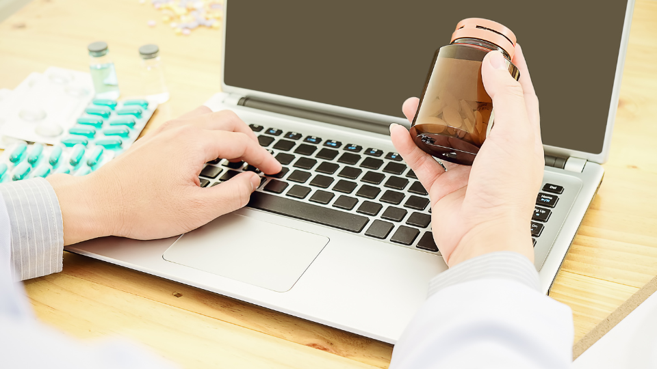 Pharmacist holding a bottle of pills and working on a laptop.