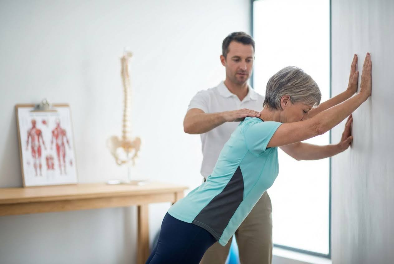 Physiotherapist guiding a woman in a teal shirt doing a wall push-up exercise, with a spine model in the background