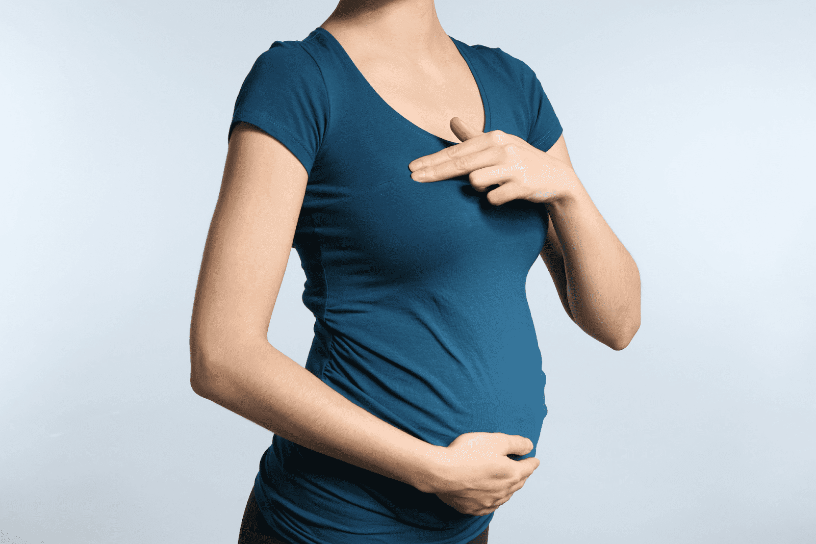 Pregnant woman in a teal shirt performing a breast self-exam, with one hand on her belly and the other pointing to her chest.