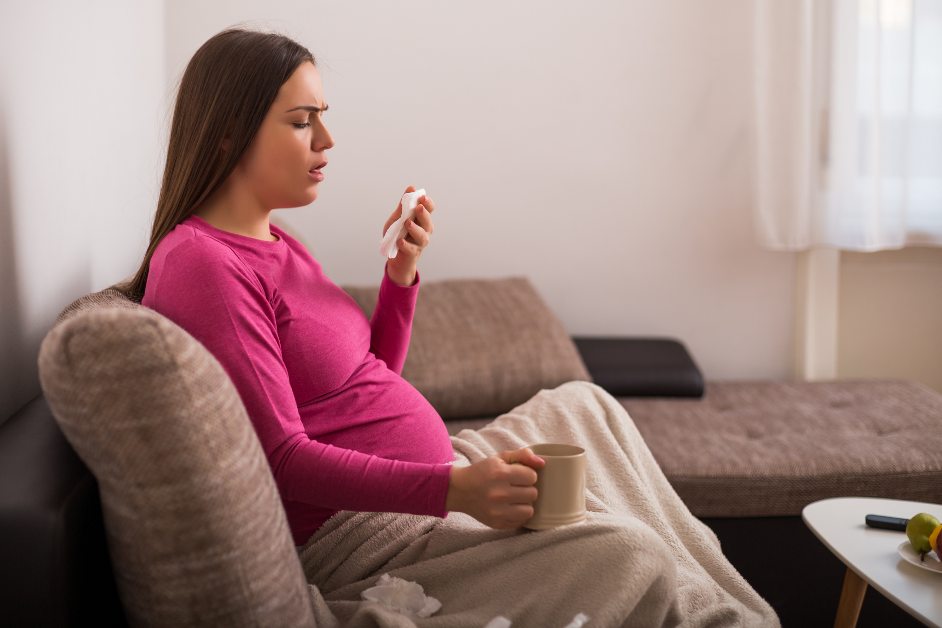 Pregnant woman sitting on a couch holding a tissue and a mug, appearing uncomfortable with a cough or sore throat