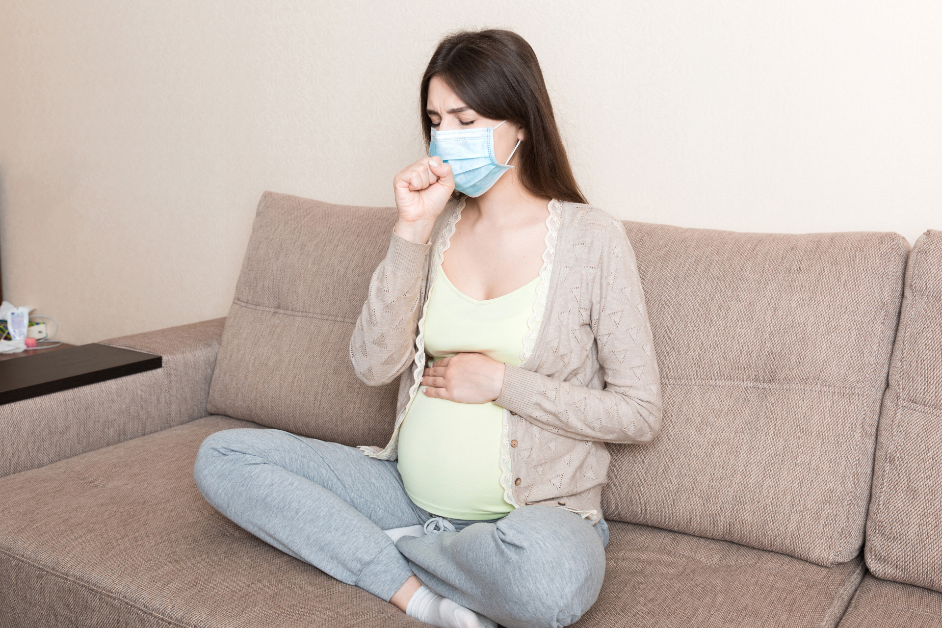 Pregnant woman wearing a face mask and coughing while sitting on a couch
