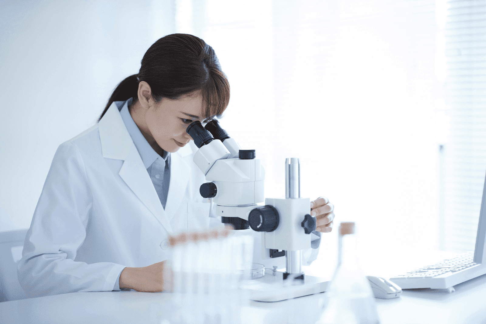 Scientist in a white lab coat using a microscope in a bright laboratory with test tubes and research equipment on the table.