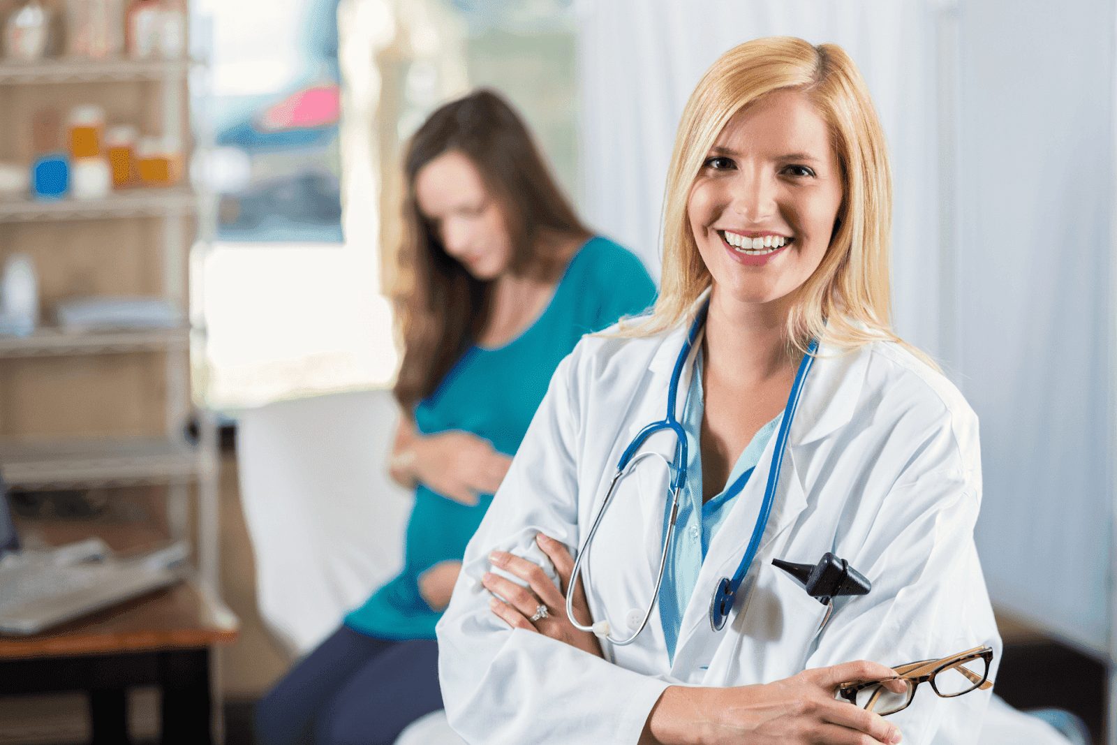 Smiling doctor in a white coat with a stethoscope, standing in front of a pregnant patient sitting in the background