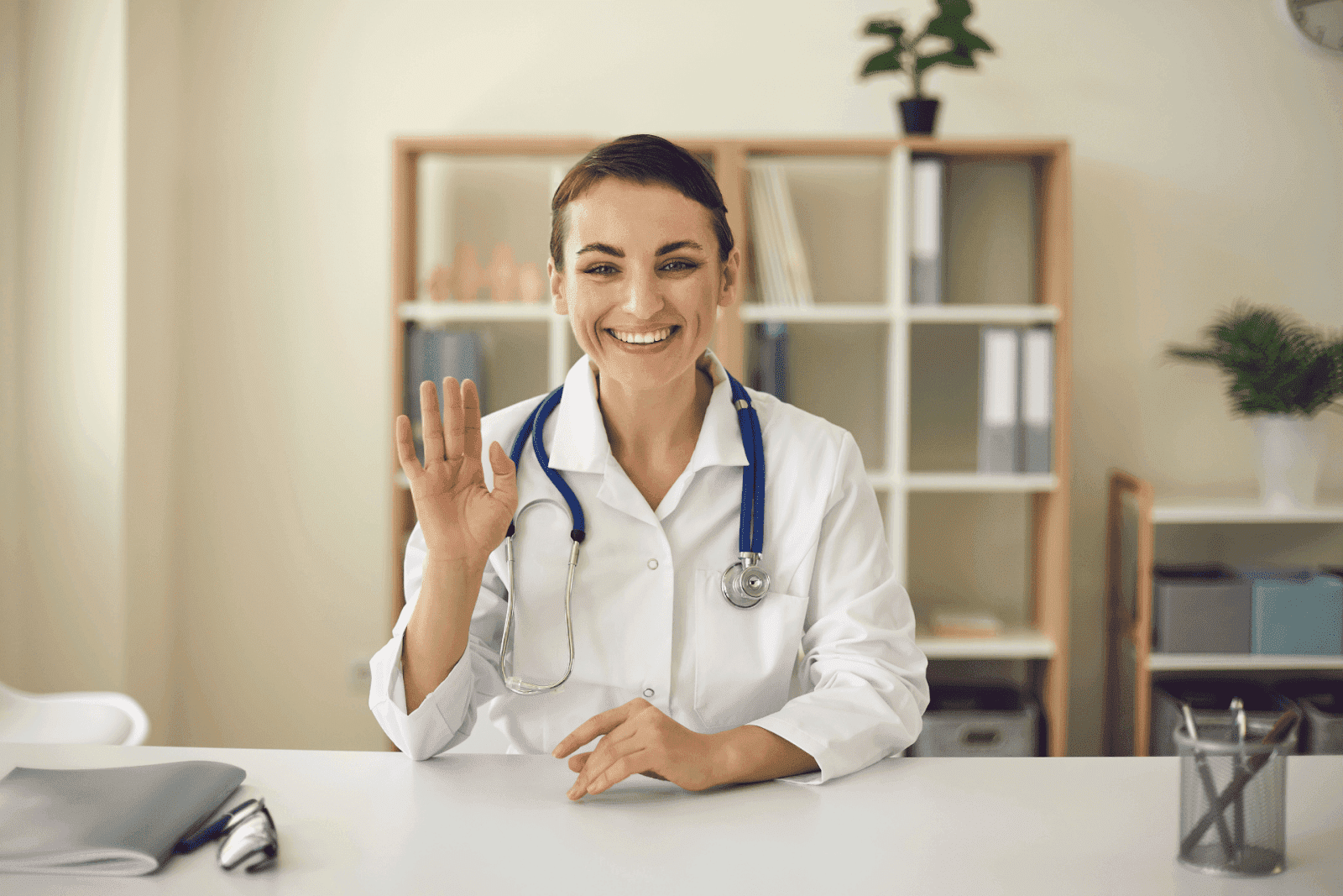 Smiling female doctor waving during a virtual consultation in a bright office.