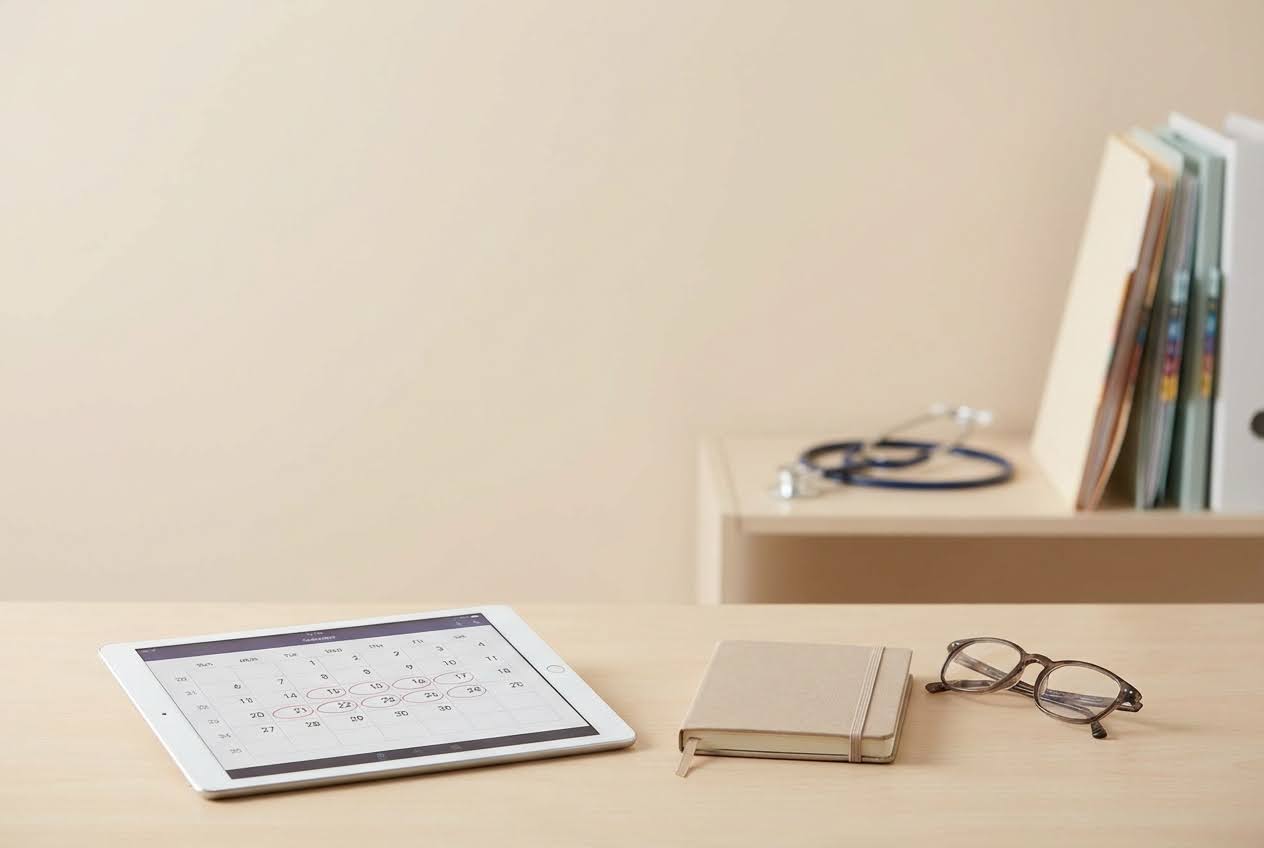 Tablet displaying a calendar with circled dates, a notebook, and glasses on a wooden desk, with a stethoscope in the background.