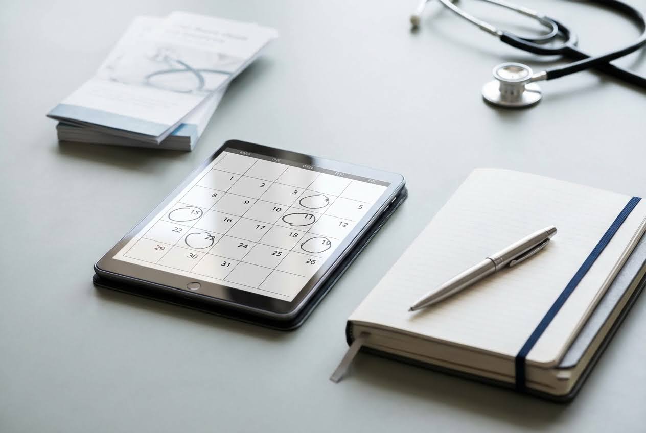 Tablet displaying a calendar with circled dates, a notebook with a pen, and a stethoscope on a light gray surface.