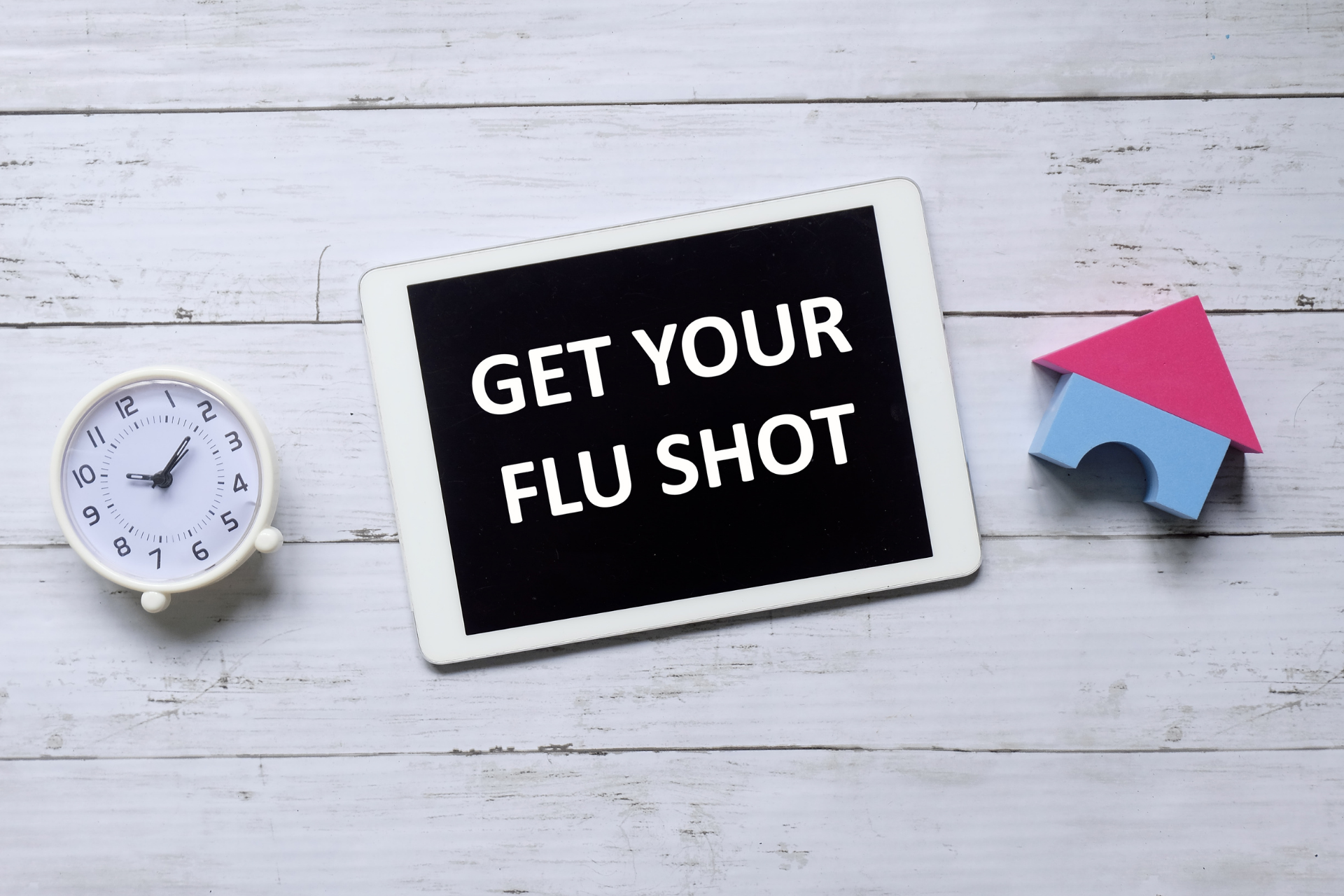 Tablet displaying the text ‘Get your flu shot’ beside a clock and a small house icon on a white surface
