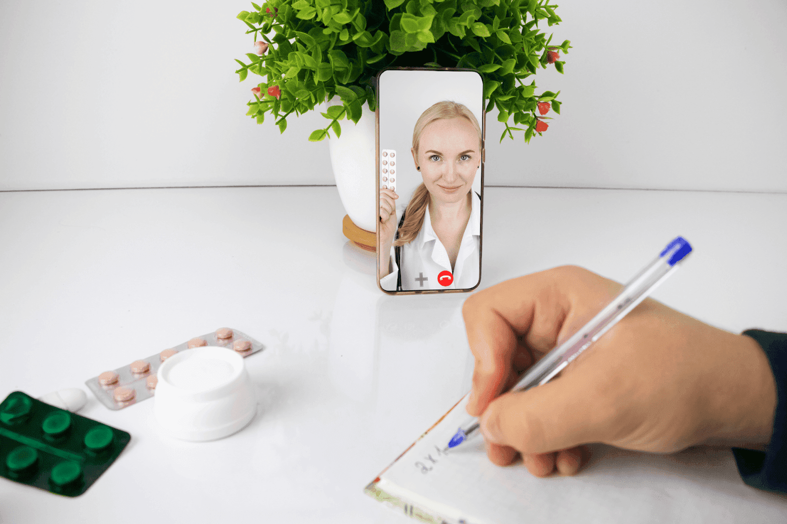 Telehealth consultation on smartphone with a female doctor showing medication while patient takes notes at home.