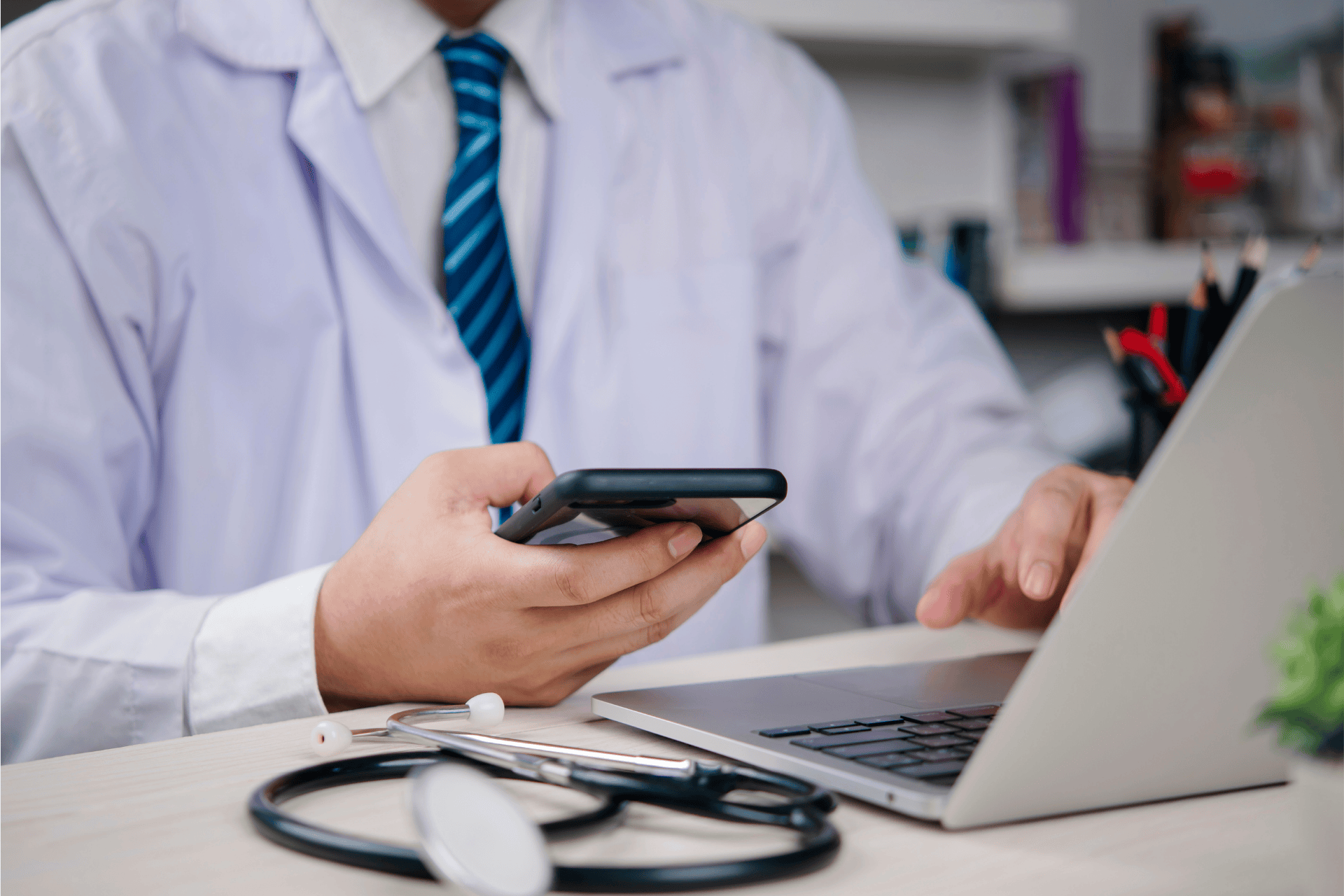 Doctor using a smartphone and laptop at a desk with a stethoscope nearby.