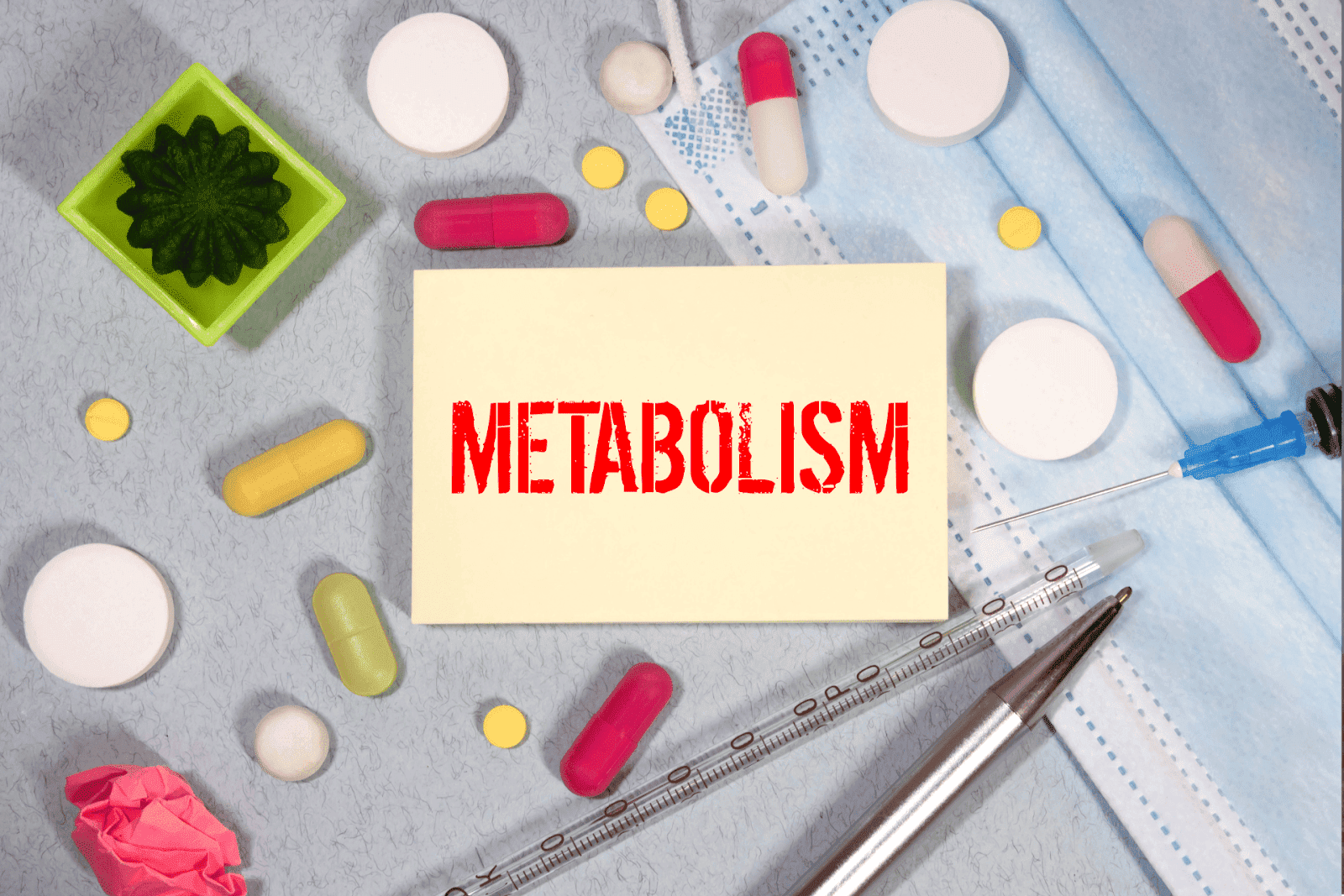 The word Metabolism written on a note surrounded by pills, a syringe, and a face mask.