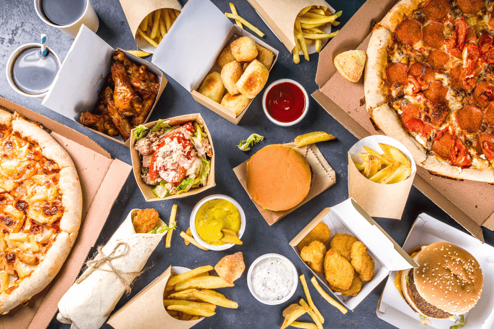 Top-down view of assorted fast food, including pizzas, burgers, fries, chicken wings, nuggets, wraps, and dipping sauces