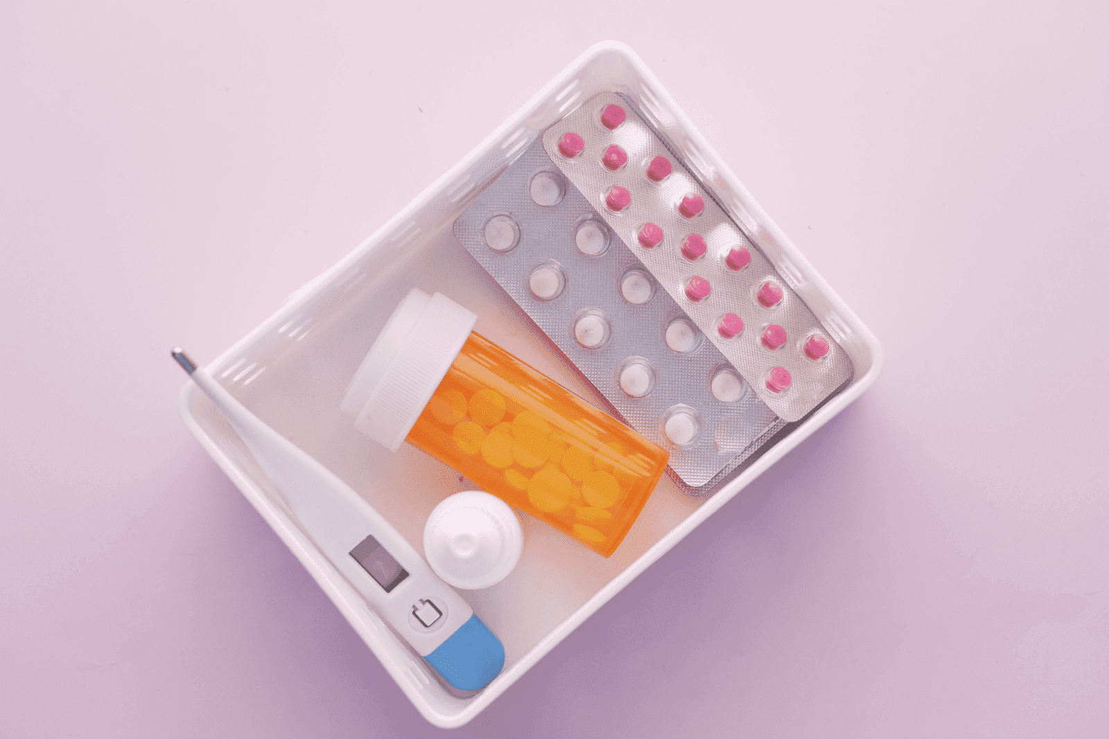 Tray with eczema pills, blister packs, a pill bottle, and a digital thermometer on a pink background.