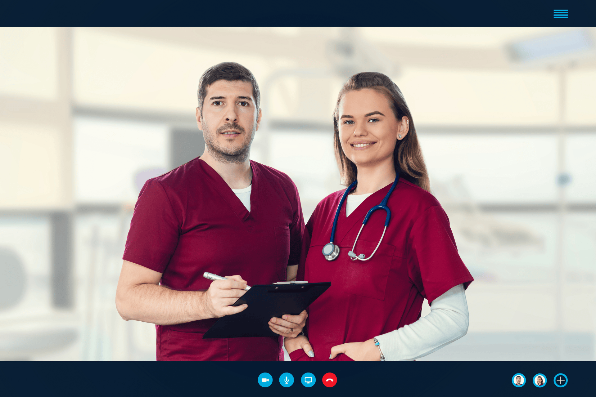 Two healthcare workers in burgundy scrubs standing together, one holding a clipboard.